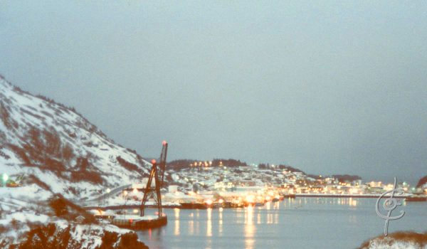 A view of Kodiak City at winter's dusk. Lights from the boat harbor burn a distant gold, appearing to melt into the bay below the braakwater.